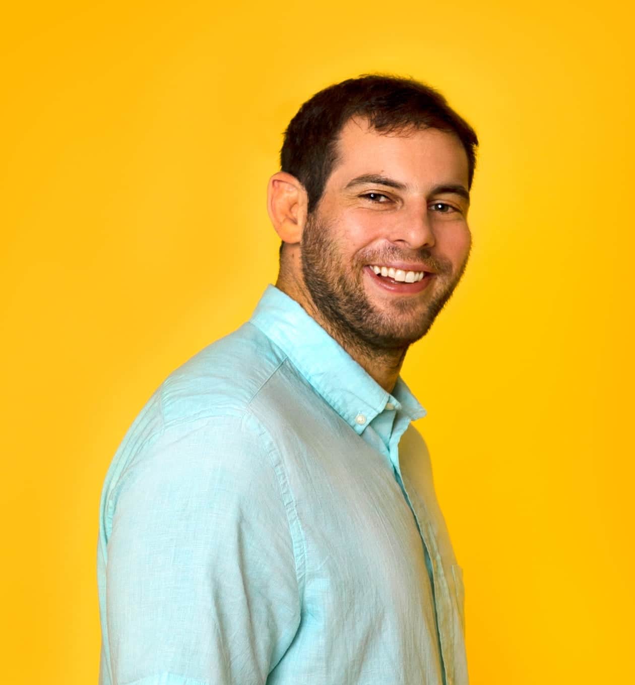 A man with short dark hair and a beard, wearing a light blue button-up shirt, smiles while standing against a bright yellow background.