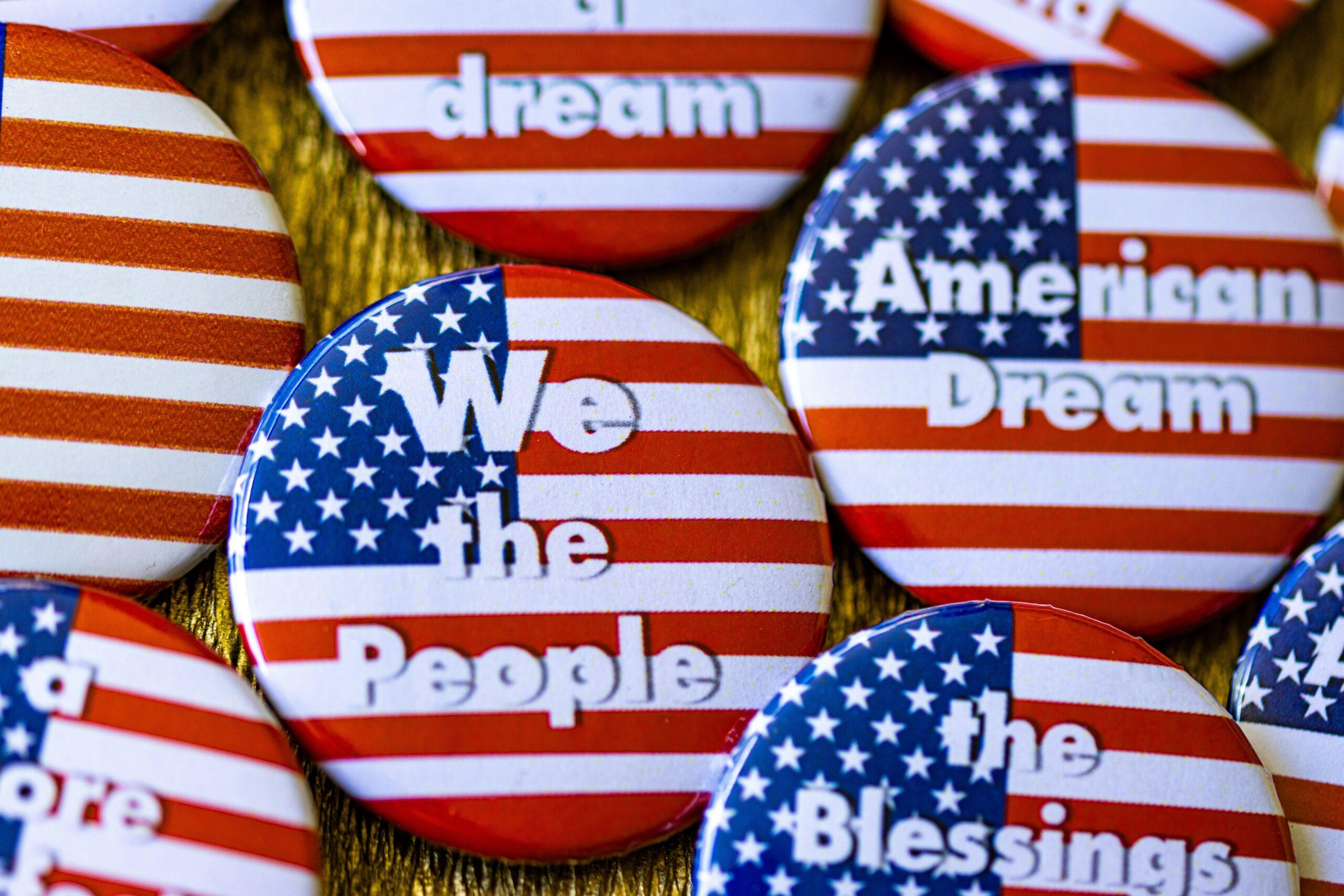 A collection of American flag pins sits on a table. Each pin has a patriotic message, like 