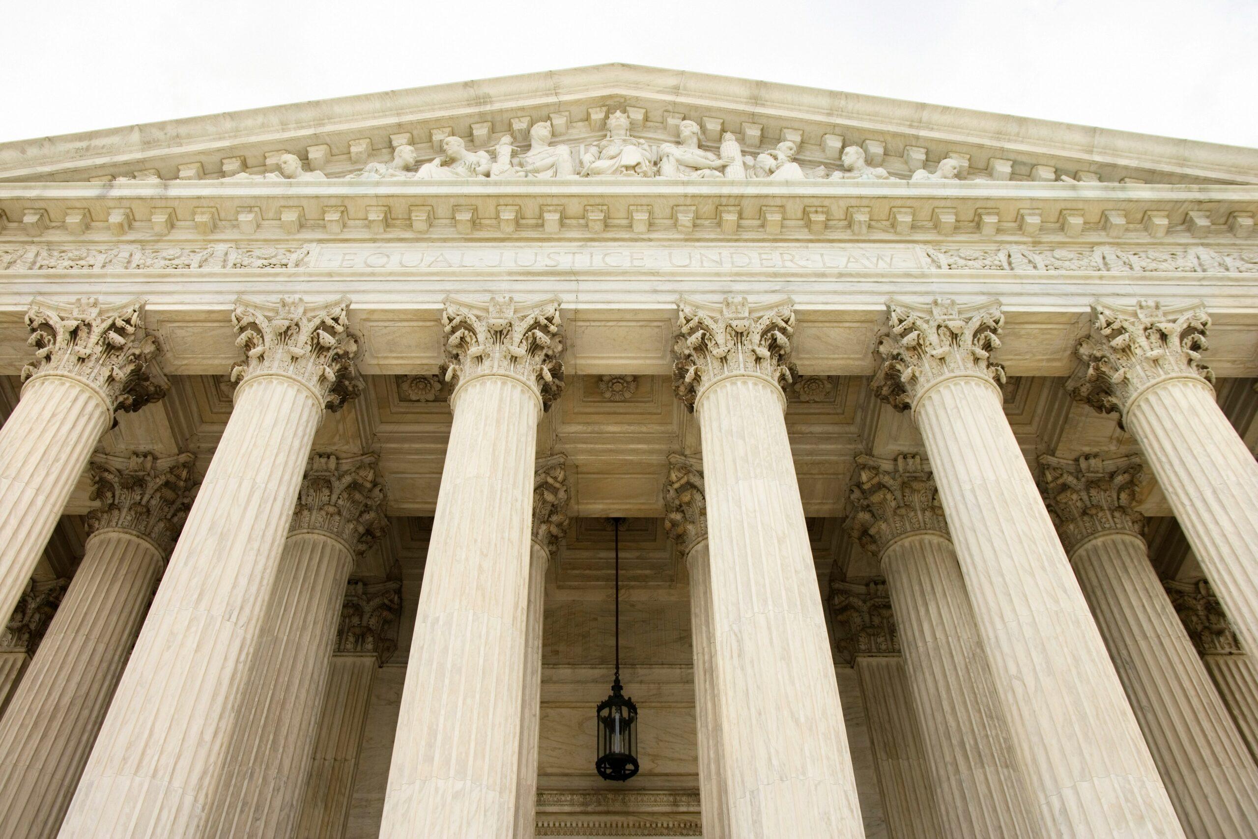 Low-angle view of a grand neoclassical courthouse façade with tall marble columns, ornate capitals, and a sculpted pediment featuring figures above the inscription “Equal Justice Under Law.”