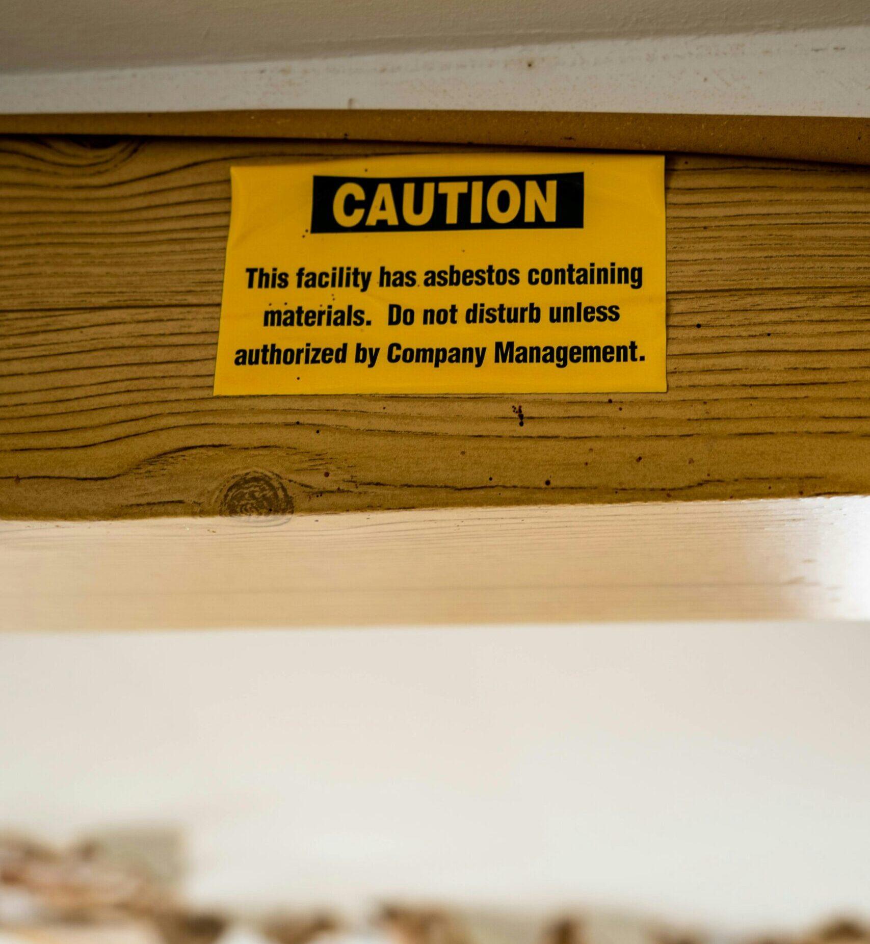 Yellow warning label attached to a wooden beam stating “Caution: This facility has asbestos containing materials. Do not disturb unless authorized by Company Management,” indicating the presence of asbestos inside the building structure.