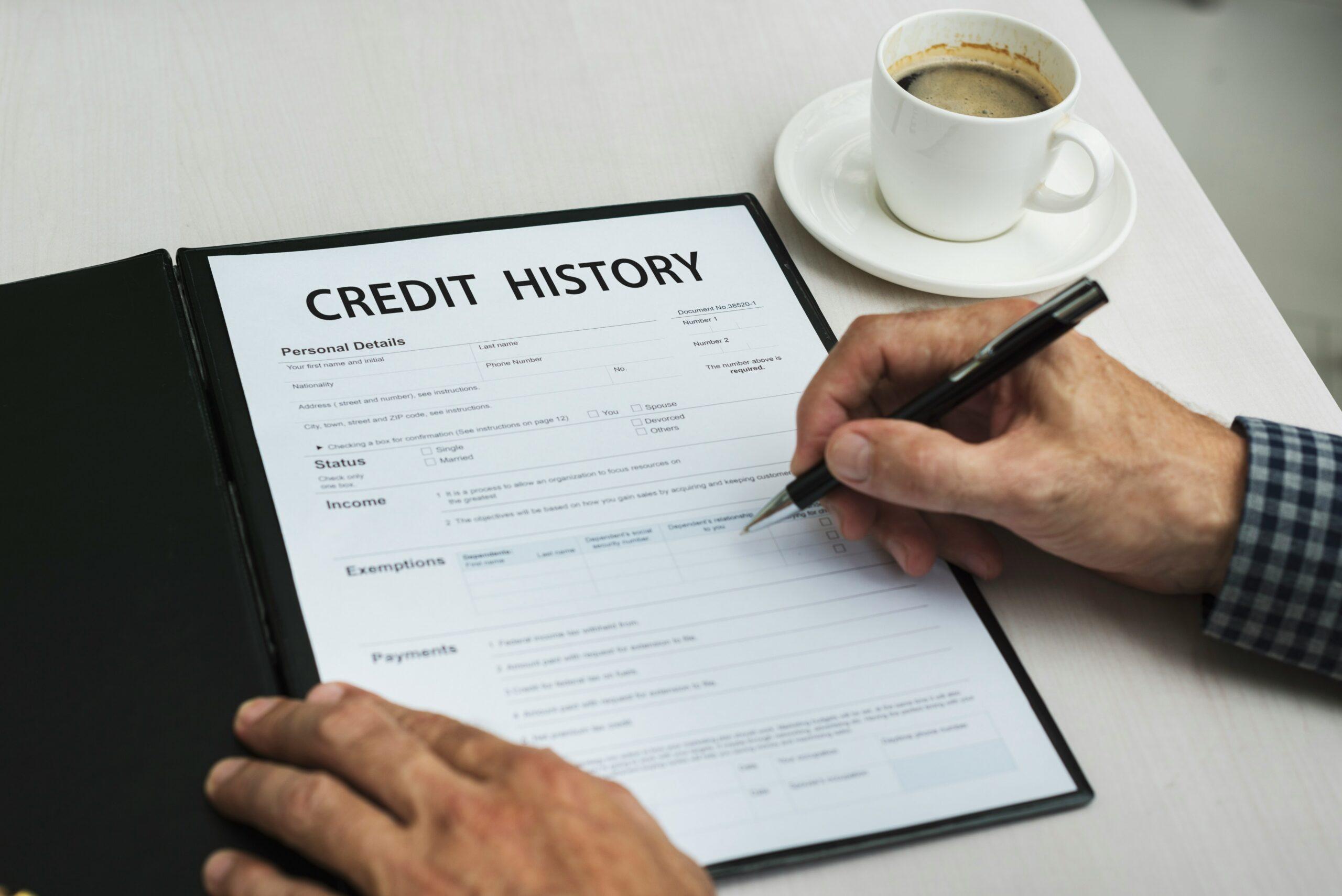 Hands holding a pen while filling out a credit history form on a clipboard, with sections for personal details, income, and payments visible, and a cup of coffee on a saucer placed on the table nearby.