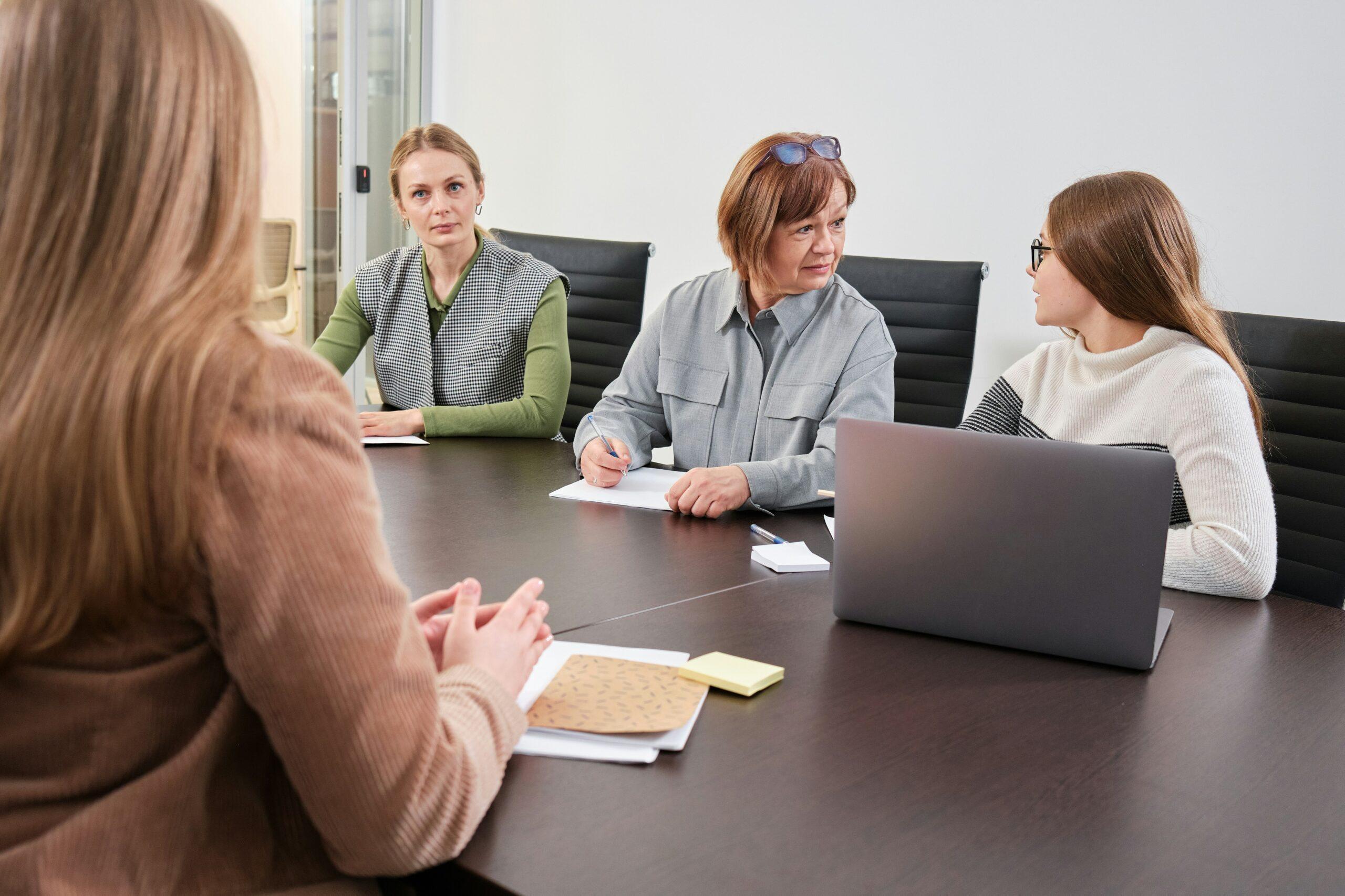 Four women seated around a modern conference table during a professional meeting, with one woman speaking in the foreground while three others listen attentively, taking notes and looking toward her; a laptop, notepad, and sticky notes are visible on the table in a bright office setting with neutral walls and black office chairs.