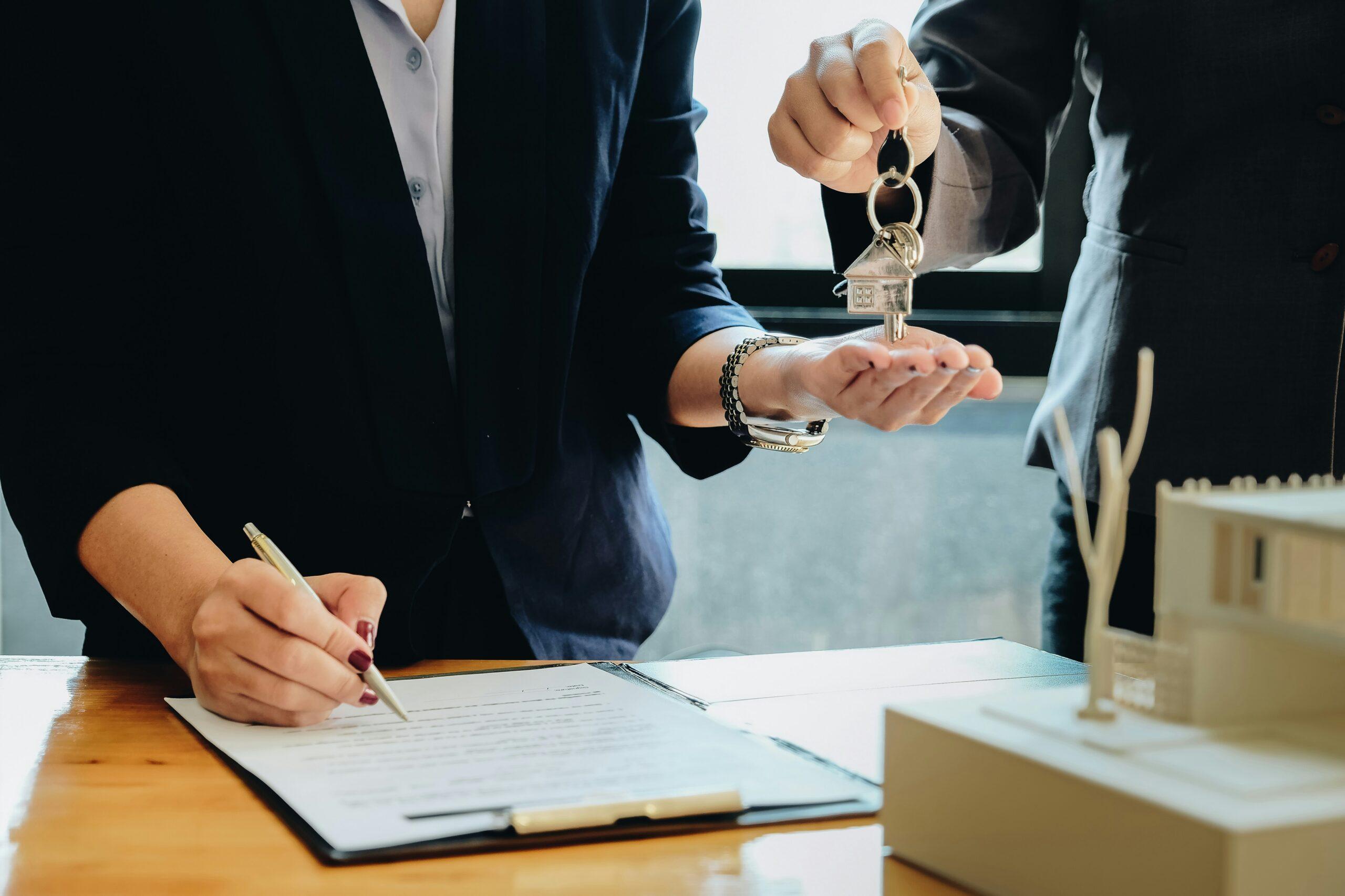 Close-up of a real estate transaction where one person signs a contract on a clipboard while another hands over a set of house keys with a small house-shaped keychain, symbolizing the transfer of property ownership.