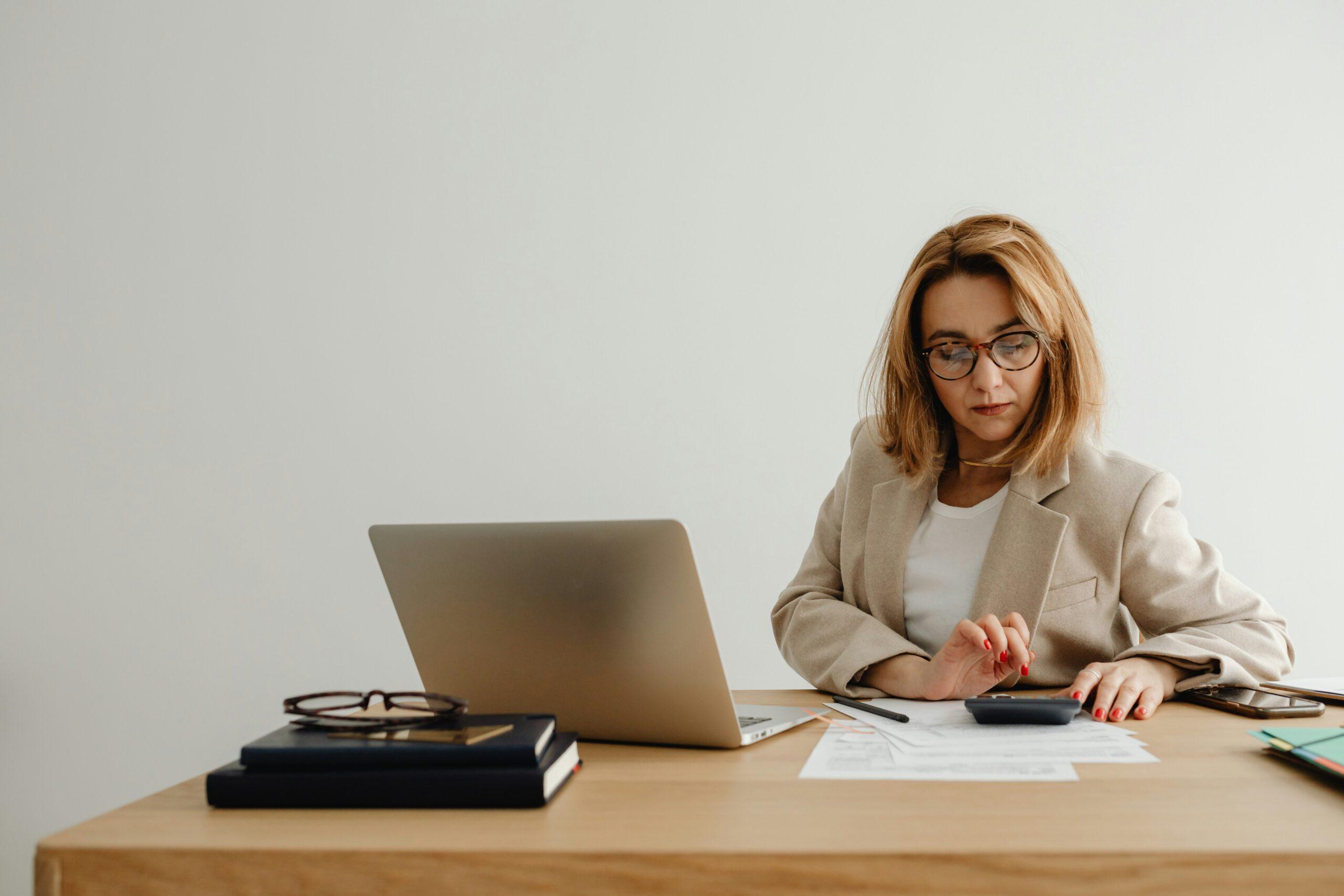 Woman wearing glasses and a beige blazer sits at a wooden desk against a plain light background, focused on reviewing printed documents while using a calculator, with an open laptop, stacked books, eyeglasses, and a smartphone placed neatly on the desk, conveying concentration, financial or administrative work, and a calm professional workspace.