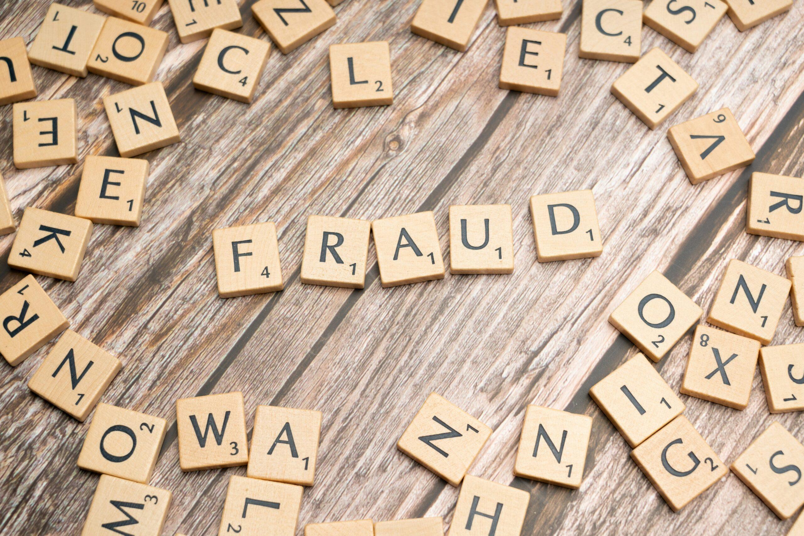 Wooden letter tiles scattered across a rustic wooden surface, with the letters arranged in the center to clearly spell “FRAUD,” while other tiles are randomly dispersed around, creating visual emphasis on the concept of fraud and a sense of disorder.